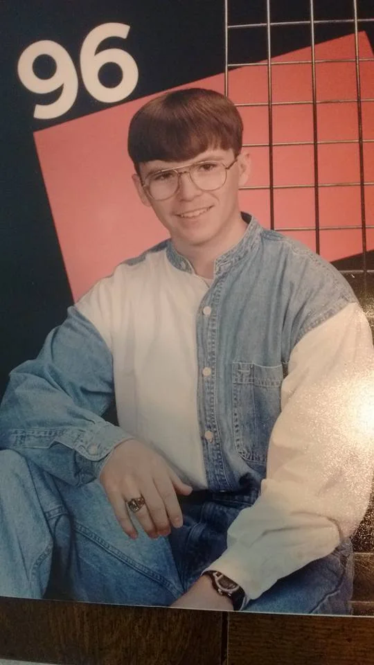 A person wearing large glasses and a denim and white shirt sits in front of a geometric background. The number "96" is in the top left corner. They are smiling with one knee raised and wearing a ring on their right hand.