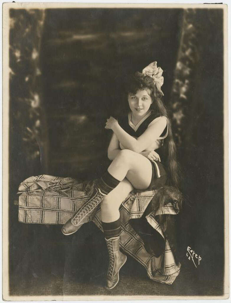 A vintage sepia-toned photograph of a woman sitting on a patterned fabric-covered box. She's wearing a short outfit with tall lace-up boots and has a large bow in her long hair, smiling slightly at the camera.