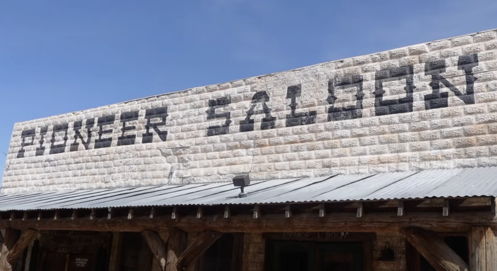 Old stone building with a wooden facade and a corrugated metal roof. The words "SINGER SALOON" are painted in large, faded black letters on the stone wall. The sky is clear and blue in the background.