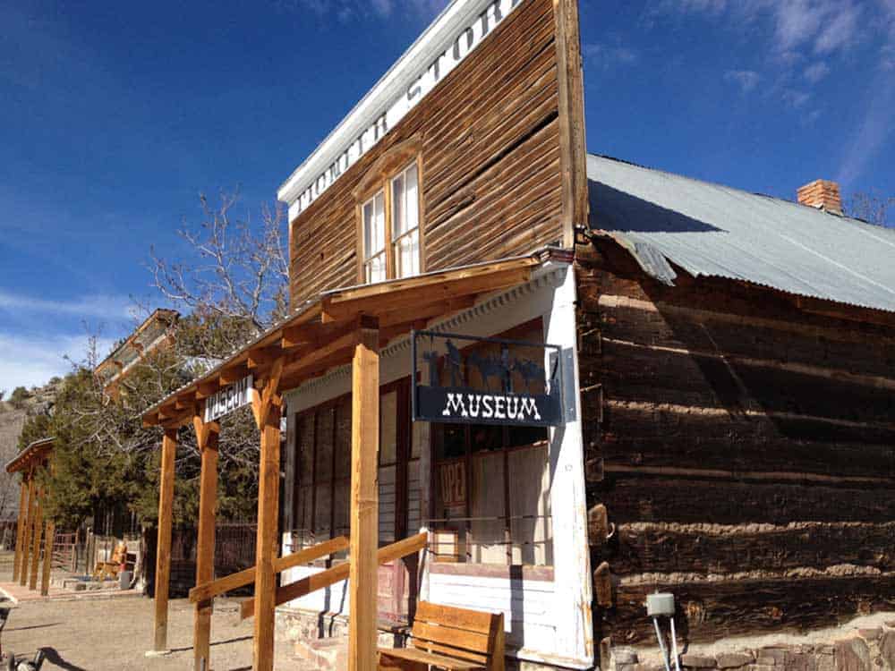 Old wooden building labeled "Museum" with a rustic porch, weathered wood siding, and a metal roof against a blue sky. Trees and a dirt path are in the background.