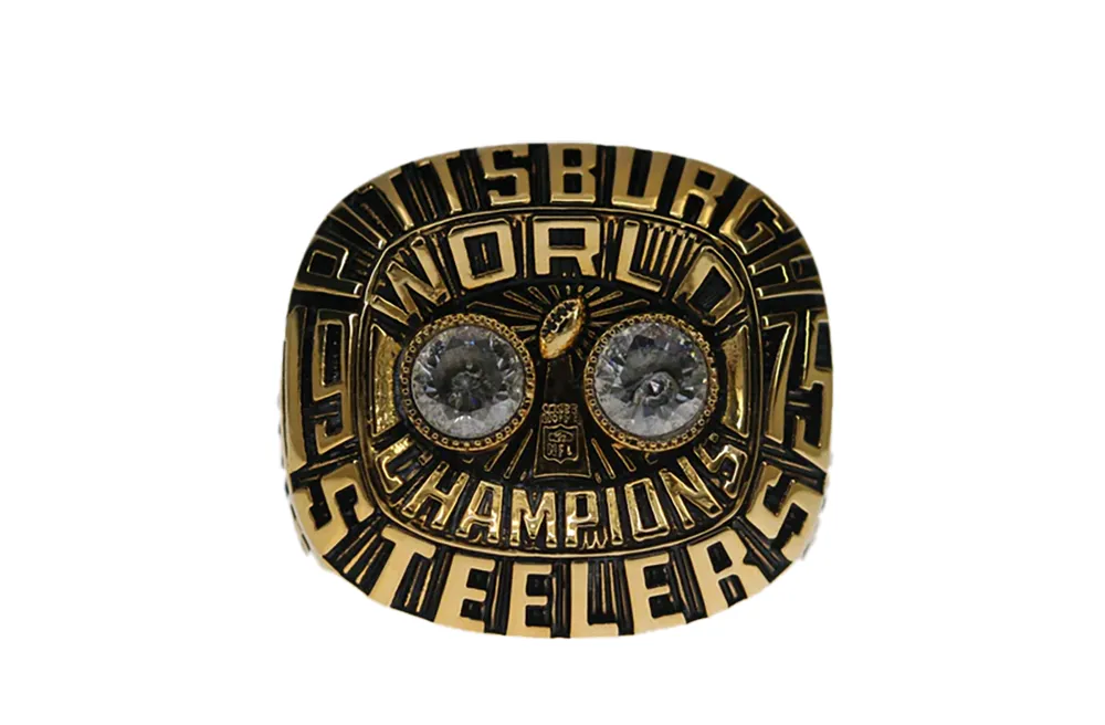 Close-up of a gold Pittsburgh Steelers championship ring featuring bold letters that read "WORLD CHAMPIONS," with two large diamond-like stones in the center.