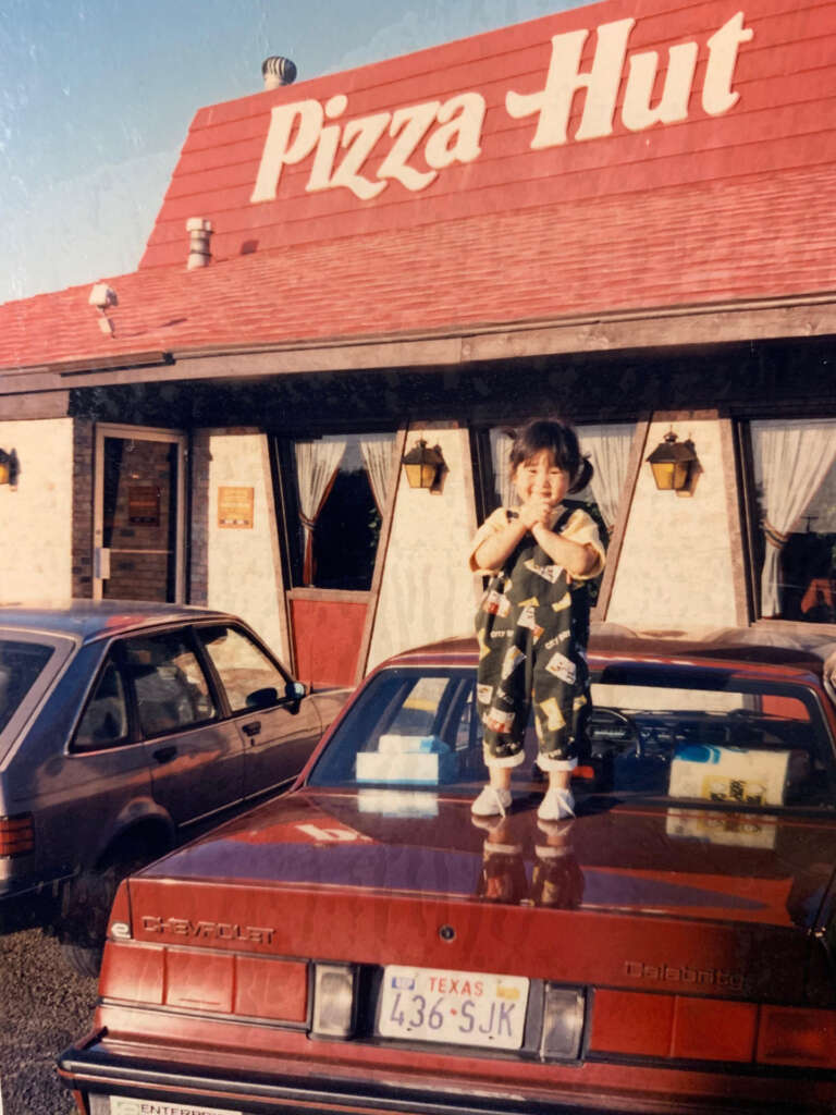 A child stands on the hood of a red car in front of a Pizza Hut building. The child is smiling and wearing patterned overalls. Two cars are parked in the lot, and the Pizza Hut sign is visible above.