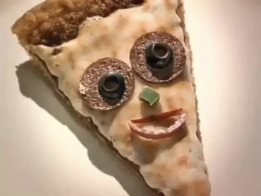 A slice of pizza with a human-like face made from toppings. Two sausage slices with black olive centers form the eyes, a small piece of green pepper for the nose, and a curved red pepper for the mouth. The background is plain white.