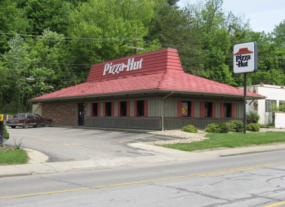 A Pizza Hut restaurant with a distinctive red roof and brick exterior is surrounded by trees. A sign with the Pizza Hut logo stands by the road. The parking lot is mostly empty, and the sky is clear.