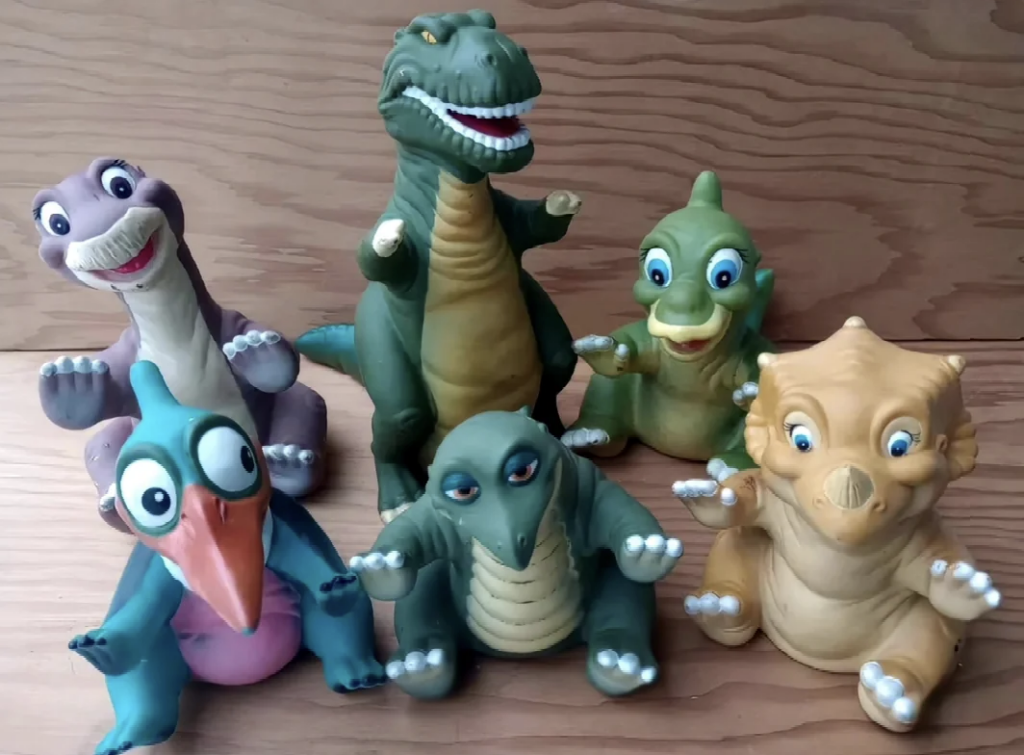 A group of six dinosaur character toys are arranged on a wooden surface. The toys are colorful and cartoonish, each with distinct features like big eyes and expressive faces. They resemble popular prehistoric animals like T-Rex, triceratops, and pterodactyl.