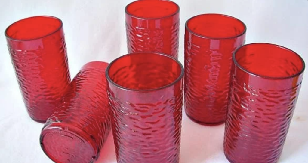 Six textured, red glass tumblers are arranged on a white surface. Five stand upright while one lies on its side, showing the base. The vibrant red hue highlights the intricate pattern on each glass.