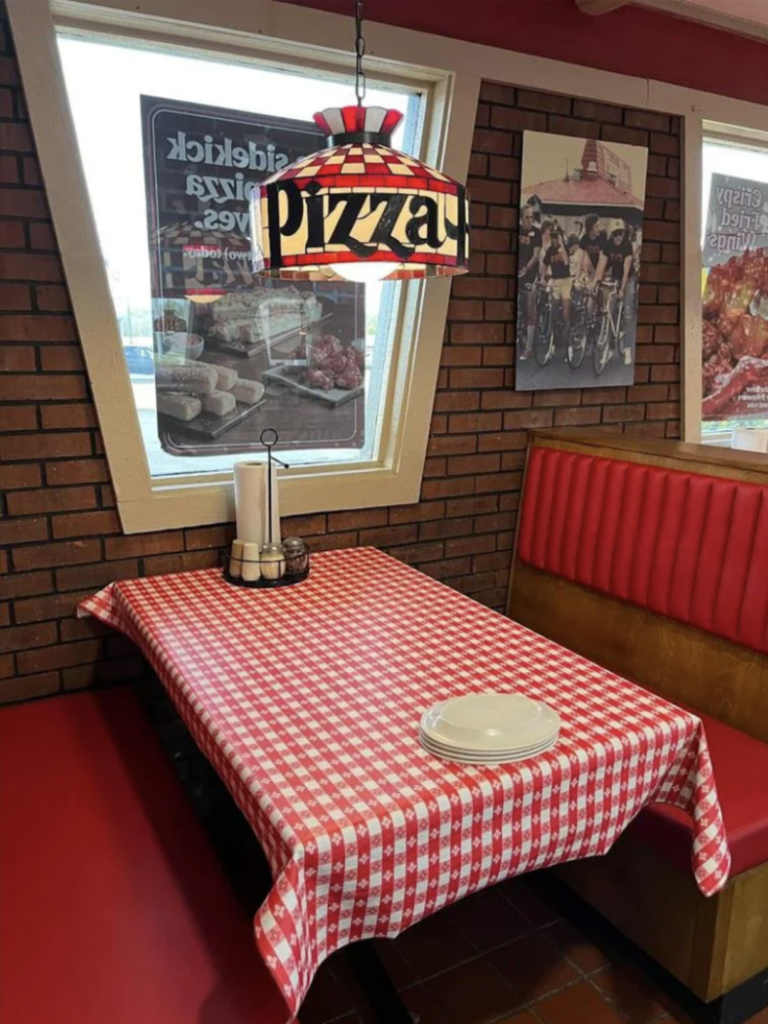 A restaurant booth with red and white checkered tablecloth, empty white plates, and napkins. A hanging lamp above has "pizza" written on it. Brick walls display posters, and red cushioned seating is visible.