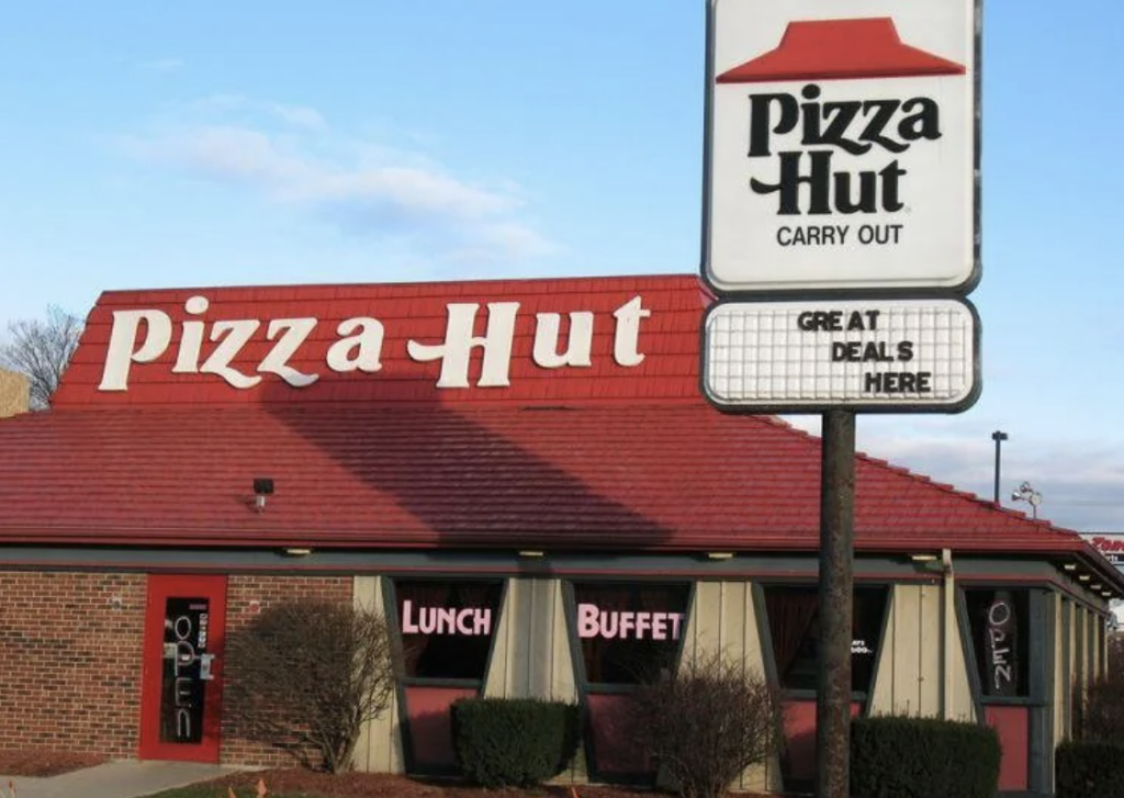 Pizza Hut restaurant with a red roof and "Pizza Hut" signage. A tall sign reads "Pizza Hut Carry Out" with a marquee below saying "GREAT DEALS HERE." The building has a "Lunch Buffet" sign and an illuminated "Open" sign on the door.