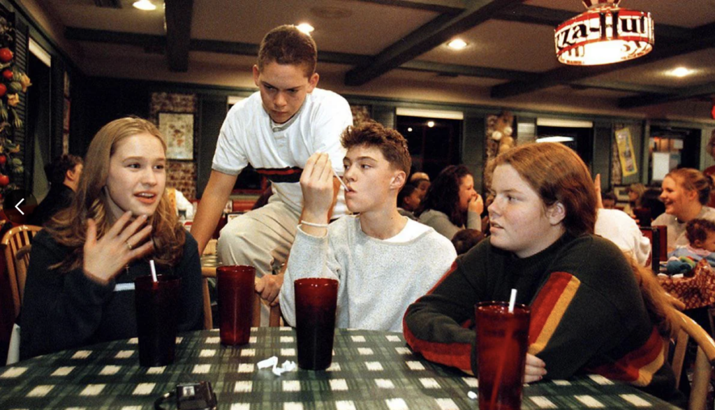 Four teenagers sit around a checkered table in a restaurant, with red drinks in front of them. One stands leaning over the table. They seem engaged in conversation. The restaurant has a casual vibe with a pizza-themed sign in the background.