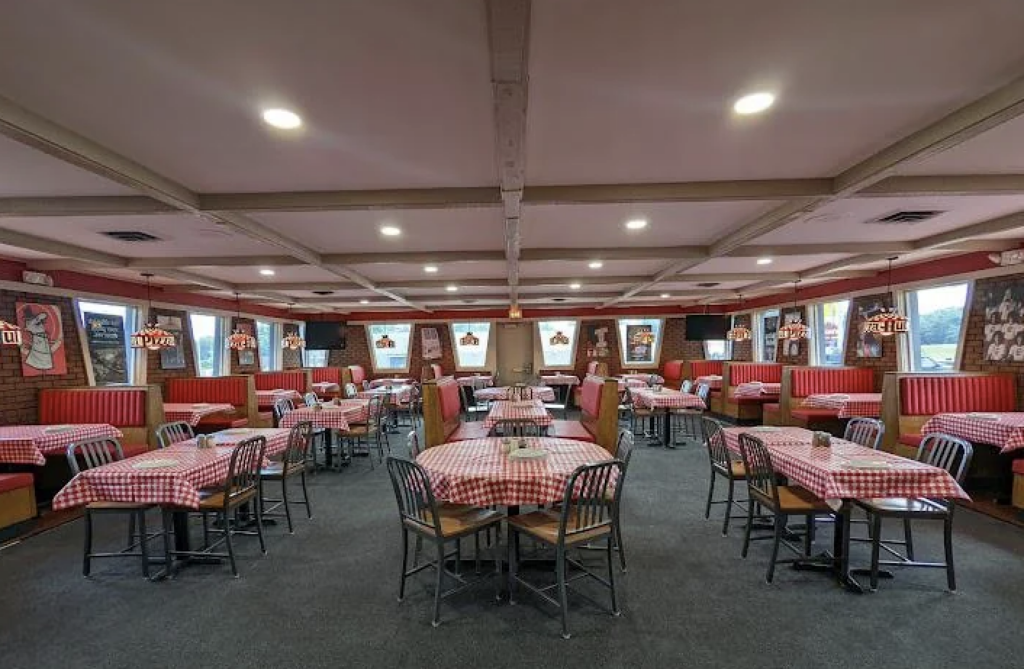 Interior of a restaurant with red checkered tablecloths, red booths, and wooden chairs. The walls are adorned with framed pictures, and the lighting is warm, creating a cozy atmosphere. The floor is carpeted, and tables are neatly arranged.