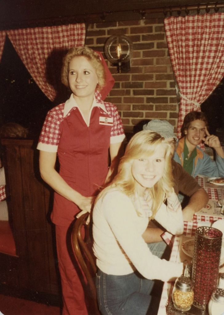 A waitress in a red and white uniform stands by a table with seated diners at a restaurant with red checkered curtains. The diners are three smiling people, two women and a man, wearing casual clothing. The atmosphere is cozy and inviting.