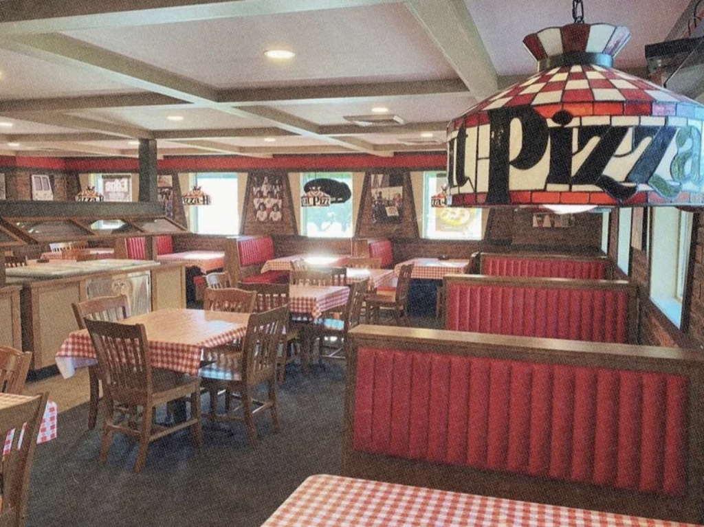 A cozy pizzeria interior with red checkered tablecloths and red booth seating. Wooden tables, chairs, and wooden-paneled walls create a warm atmosphere. A stained glass lamp with "Pizza" hangs prominently in the foreground.