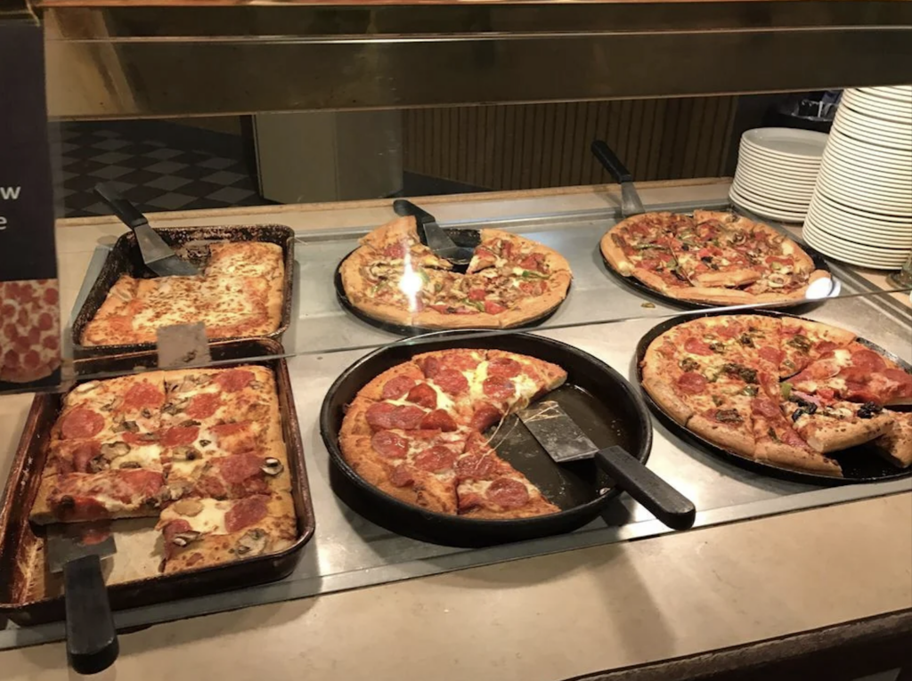 A pizza buffet featuring various types of pizzas: a rectangular cheese pizza, a pepperoni pizza, a pizza topped with vegetables and meats, and another with various toppings. Each pizza is partially sliced and served with spatulas. Plates are stacked nearby.