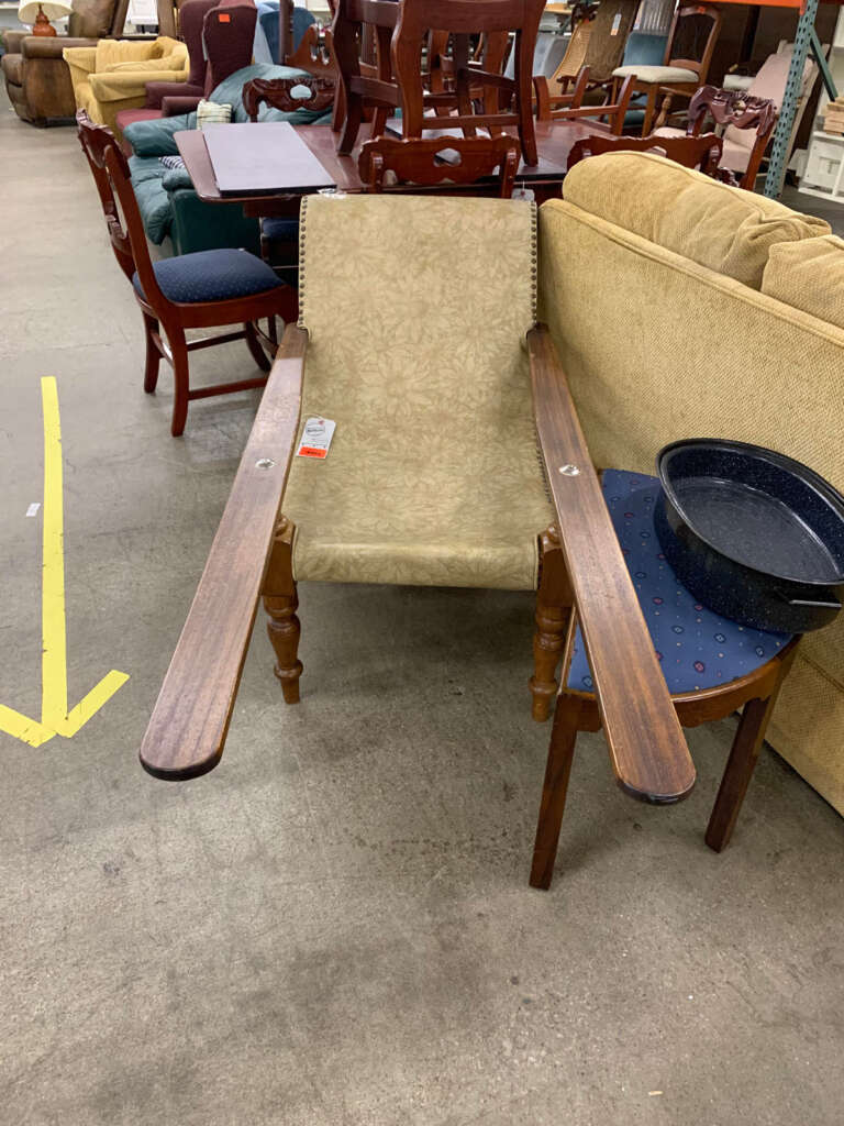 A vintage armchair with long wooden armrests and a beige patterned cushion sits in a warehouse among a variety of other furniture items. A metal bowl rests on the armchair. An arrow is painted on the floor.