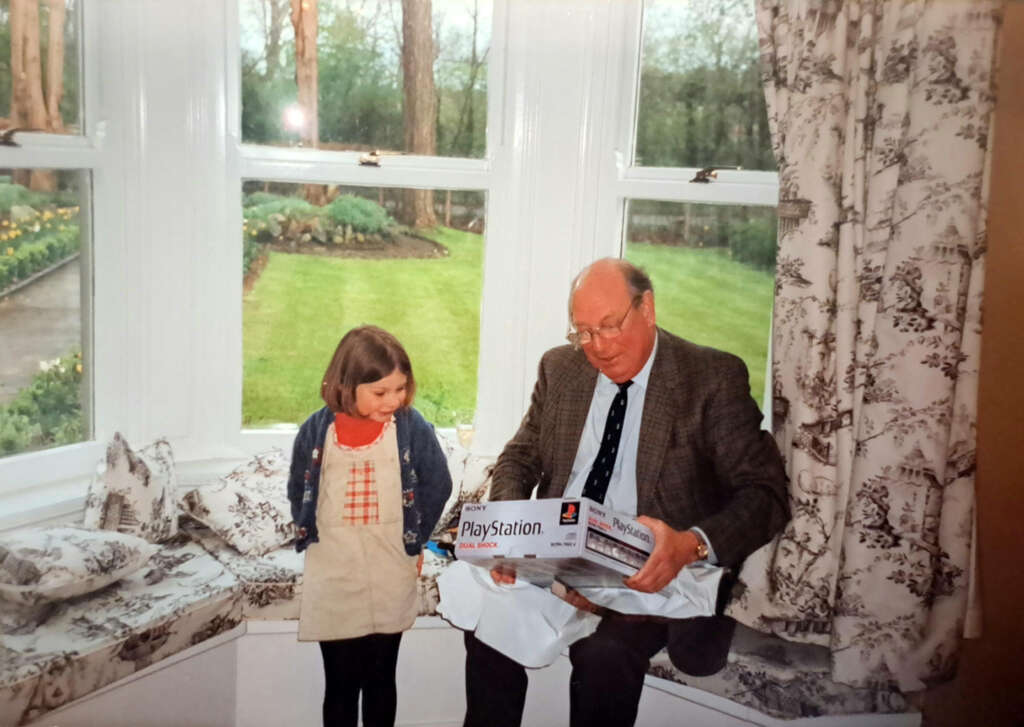 A young girl stands next to an older man sitting on a window seat. The man is holding a PlayStation console box, appearing surprised. The room has large windows with a view of a garden. Both are smiling, creating a warm, joyful scene.