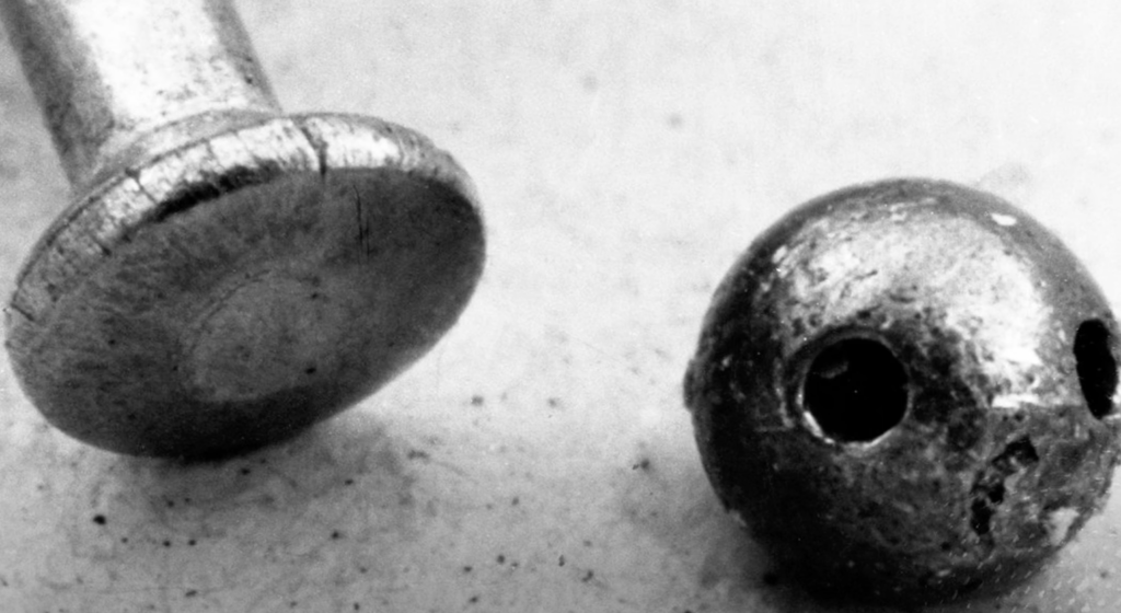 Close-up black and white image of a metal rivet next to a small, round, holed lead ball. The rivet is flattened at one end, while the ball appears to have a rough texture and two visible holes. Both objects are lying on a flat surface.