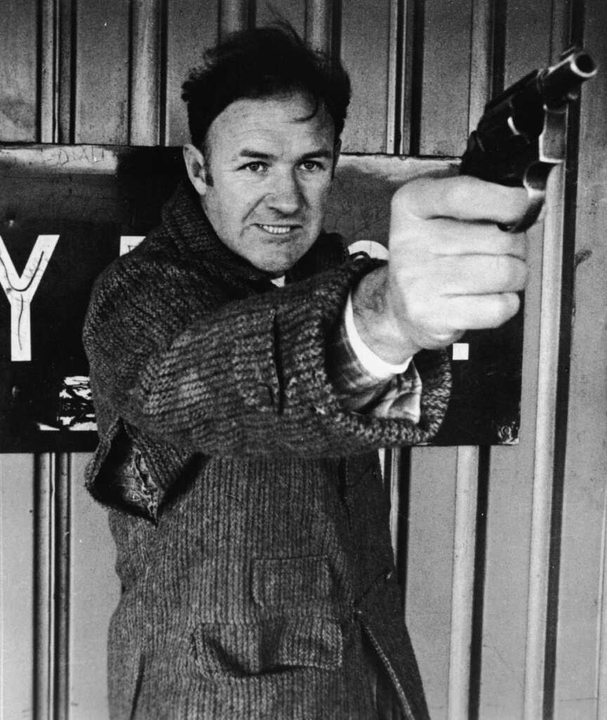 A person in a coat points a revolver directly towards the camera with an intense expression. They are standing in front of a sign with partially visible text. The image is in black and white.