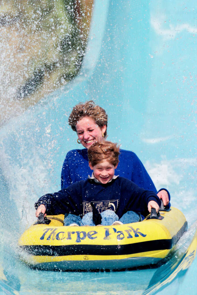 Two people, a woman and a child, ride a yellow inflatable raft down a water slide at Thorpe Park. Both are smiling with excitement as water splashes up around them. The slide is light blue, creating a dynamic, playful scene.