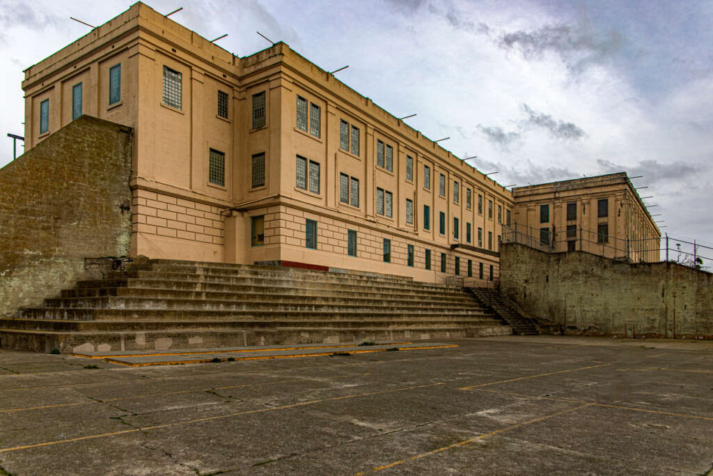 A large, weathered, beige concrete building with barred windows stands against a cloudy sky. The building is surrounded by a high wall with barbed wire. In the foreground, there are tiered concrete steps and an open, empty paved area.