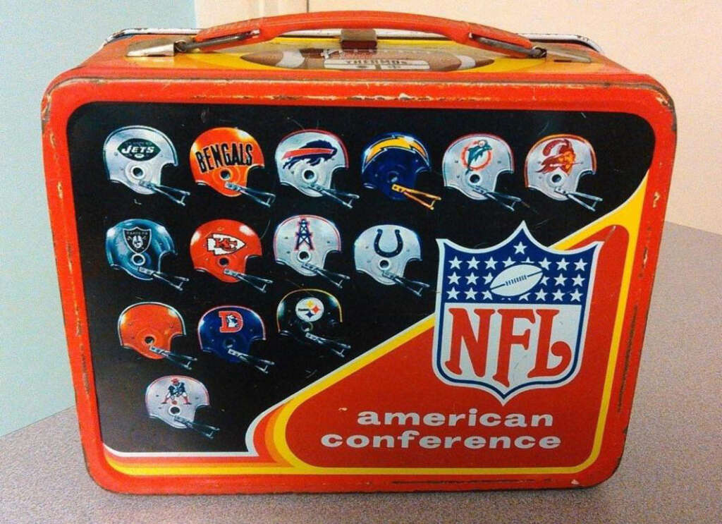 A vintage metal lunchbox featuring logos of American Conference NFL teams. The design includes helmets from various teams, arranged in rows above an NFL logo and the words "American Conference" on a red and yellow background.