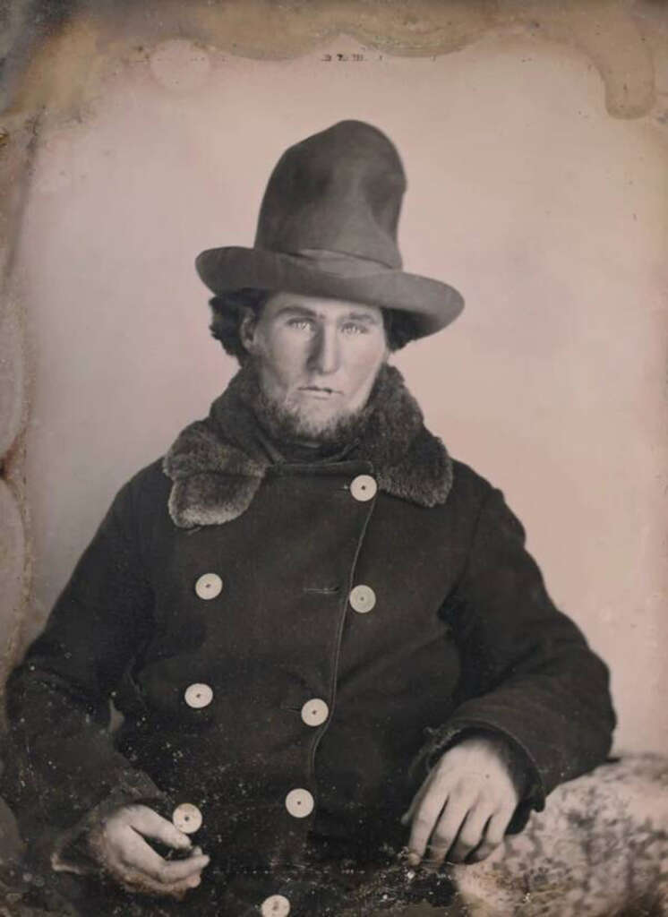 A sepia-toned photograph of a 19th-century man wearing a tall, wide-brimmed hat and a coat with large buttons. He has a serious expression and sits with his hand resting on his knee. The portrait has a vintage, weathered appearance.