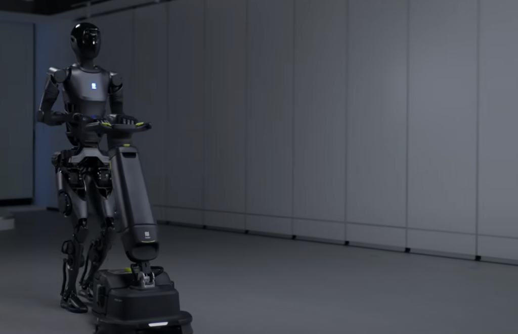 A humanoid robot is operating a floor-cleaning machine in a modern, empty room with gray flooring and white paneled walls. The robot is black and sleek, appearing to perform its task autonomously.