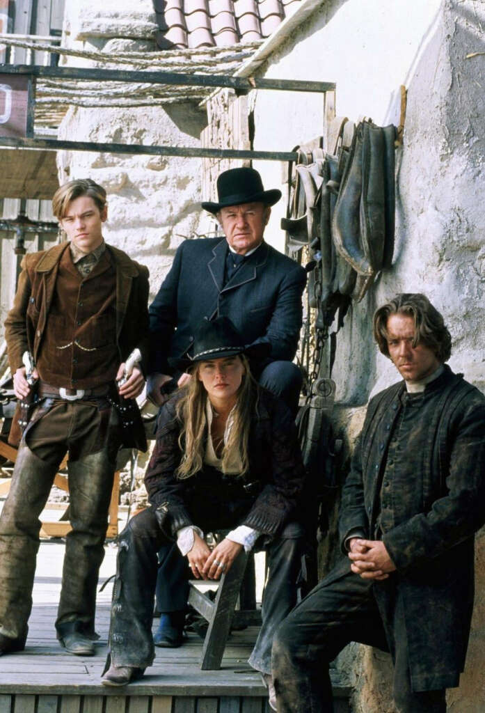 Four people in Western attire pose in an old-time setting. They wear hats and long coats, standing and sitting against a rustic, worn backdrop with old stone walls and a wooden walkway. The scene evokes a classic Western film atmosphere.