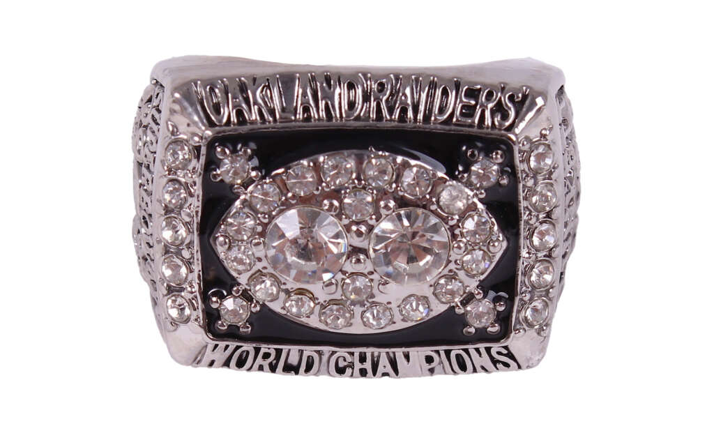 A silver ring with "Oakland Raiders" and "World Champions" inscriptions. The face of the ring features two large central stones surrounded by smaller stones in a football shape, set on a black background, with additional stones around the edges.