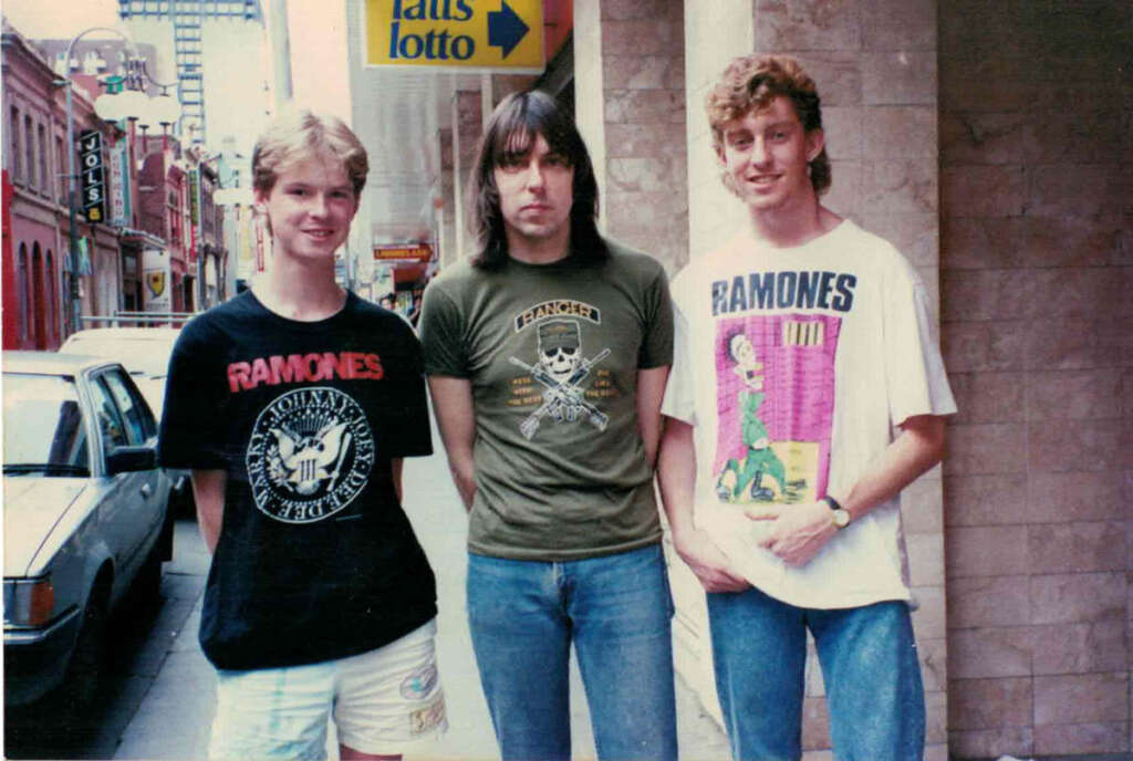 Three people stand on a city sidewalk. The middle person wears an olive graphic tee, while the others wear Ramones-themed shirts. A parked car and buildings are in the background. The "Tatts lotto" sign is visible above them.