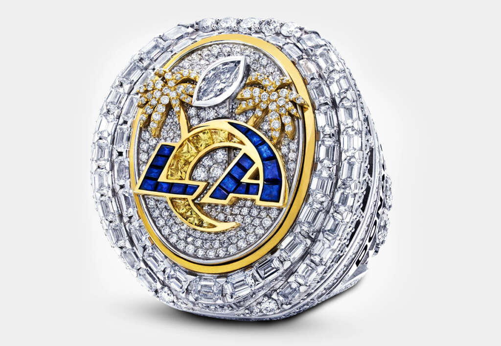 A diamond-studded ring featuring a mix of gold, blue, and silver elements. The design includes palm trees, a large "LA" in blue and gold, and a football shaped gem, symbolizing a sports championship. The ring is detailed and ornate.