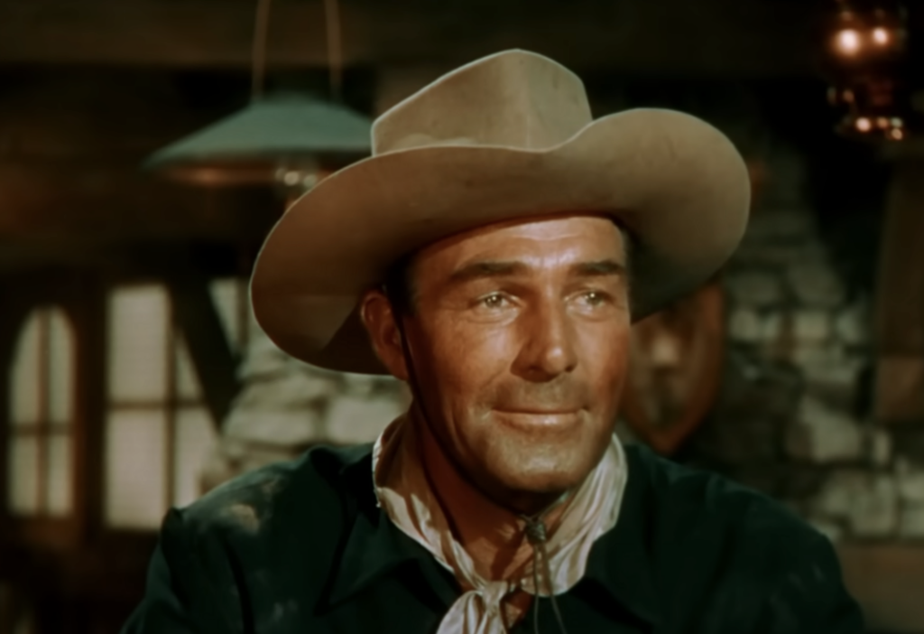 A man wearing a cowboy hat and a bandana around his neck smiles while seated indoors. The background shows a stone wall and a lamp, creating an old Western setting.