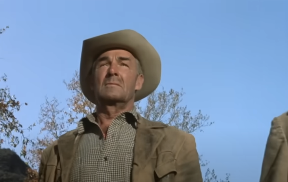 A man wearing a beige cowboy hat and brown jacket stands outdoors against a clear blue sky, looking into the distance with a serious expression. Trees with autumn leaves are visible in the background.