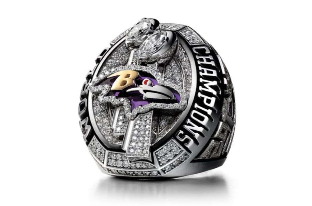 A championship ring featuring the Baltimore Ravens logo adorned with diamonds and the word "CHAMPIONS" along the side. The ring is predominantly silver with intricate detailing throughout and two large stones on top.