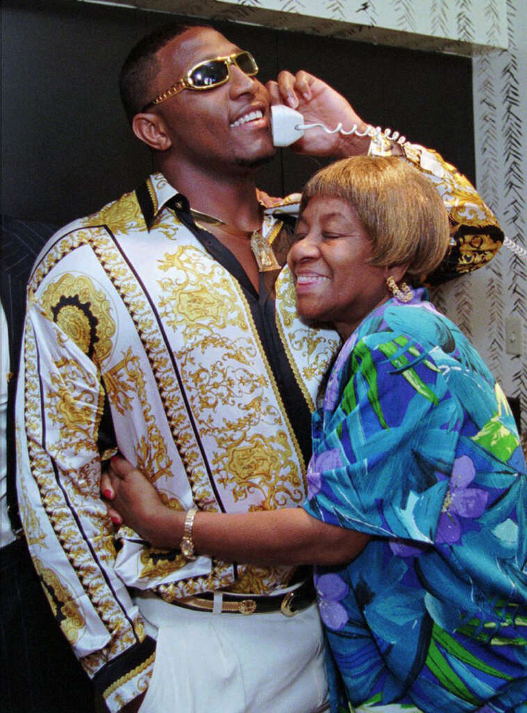 A man wearing sunglasses and a patterned shirt, holds a phone to his ear with a smile. An older woman with short hair and a colorful dress hugs him warmly, smiling with her eyes closed. They are indoors, standing closely together.