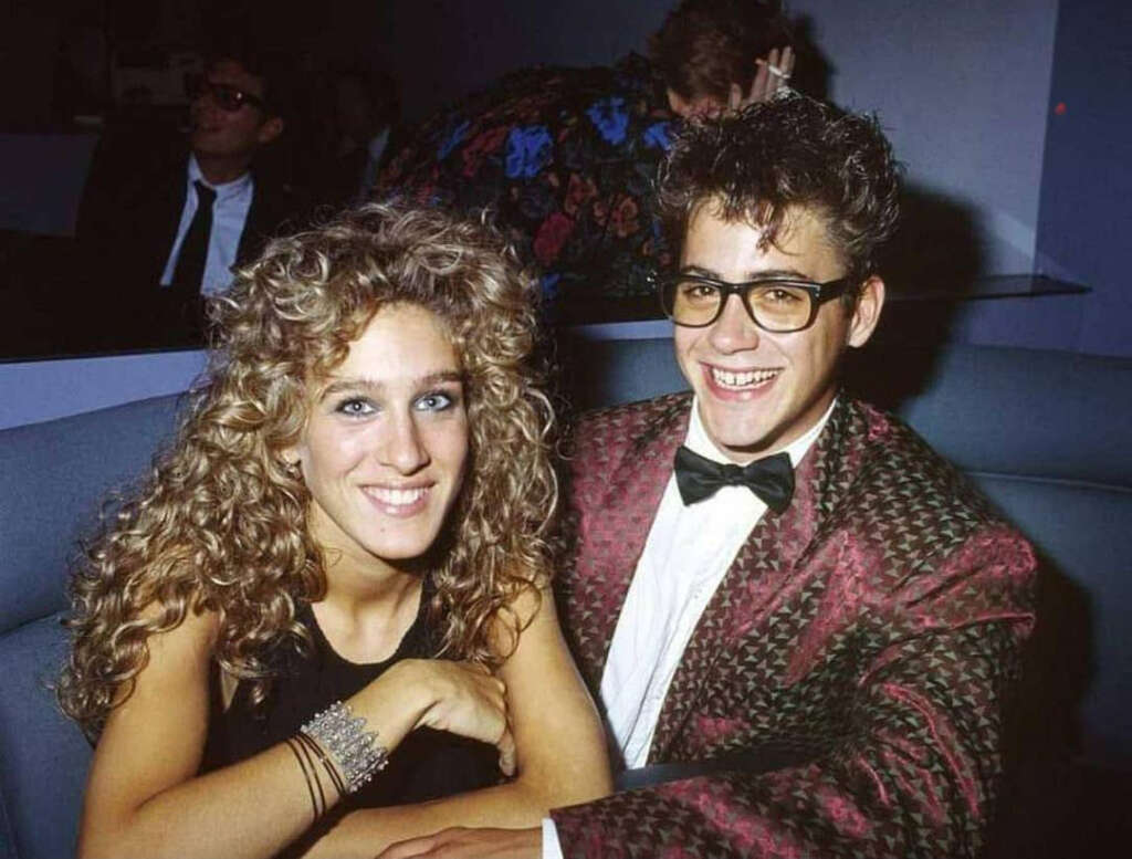 A woman with curly hair and a sleeveless top smiles while sitting next to a man wearing glasses and a patterned suit jacket. They are seated in a booth, with other people in the background. Both appear to be enjoying themselves.