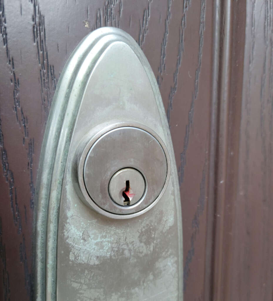 Close-up of a silver door lock with a keyhole on a dark wooden door. The lock has an oval shape, with visible scratches and signs of wear. The wood grain of the door is prominent in the background.