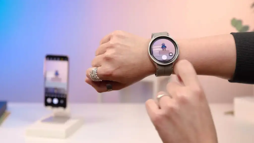 Person using a smartwatch on their wrist with a visible image on the screen. A smartphone is positioned in the background on a stand. The setting appears to be a tech-friendly environment.