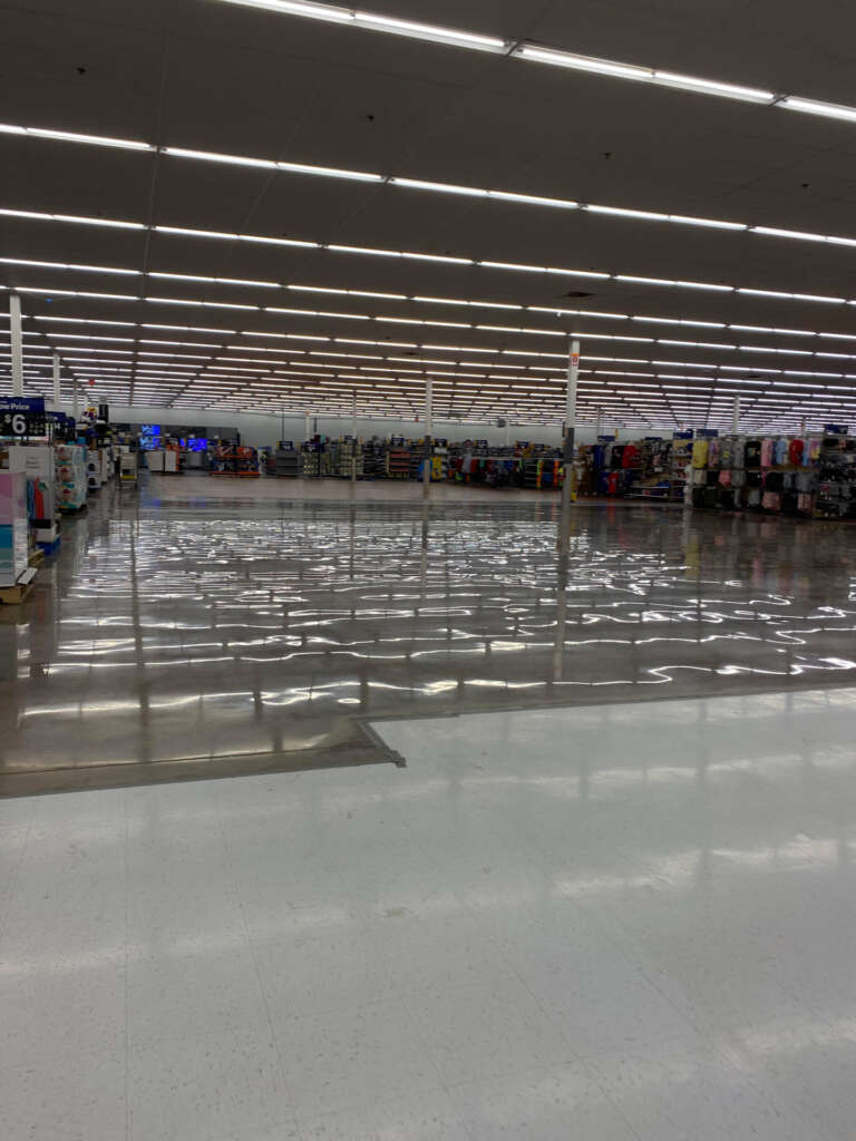 A supermarket interior with a reflective, wet floor under bright ceiling lights. Shelves are visible in the background, displaying various products. The area appears spacious and empty of people.