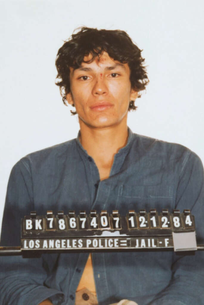 A person with curly hair and wearing a blue shirt is posing for a mugshot. The numbered sign in front reads "Los Angeles Police" with identification numbers. The person appears serious against a plain background.