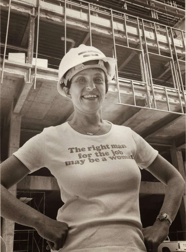 A woman wearing a hard hat and a T-shirt with the text "The right man for the job may be a woman" stands confidently in front of a construction site. The building is partially completed, with exposed concrete and scaffoldings visible.