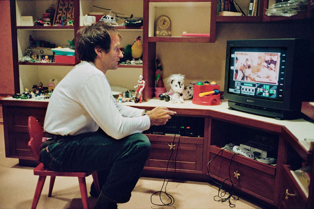 A person is sitting on a small chair, playing a video game on a CRT television. The room has shelves filled with toys, figurines, and books. The person is focused on the screen, holding a game controller. The decor is colorful and playful.