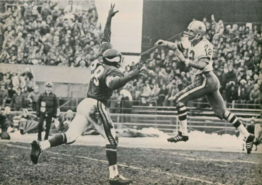 A football player in a light uniform jumps to throw a pass while a defender in a dark uniform leaps with arms raised. The crowd watches from the stands on a chilly day.