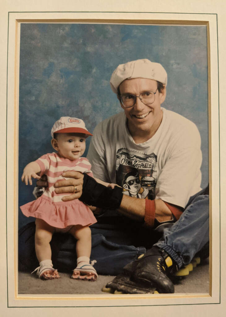 A man wearing glasses, a hat, and a Led Zeppelin t-shirt smiles at the camera while sitting on the floor. Beside him is a baby in a pink dress and cap, also looking at the camera. The background is a textured blue.