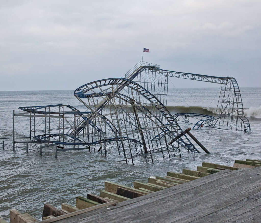 A roller coaster sits partially submerged in the ocean off a wooden pier. The structure is metal with blue tracks and a small American flag atop. Waves crash against it under a cloudy sky.