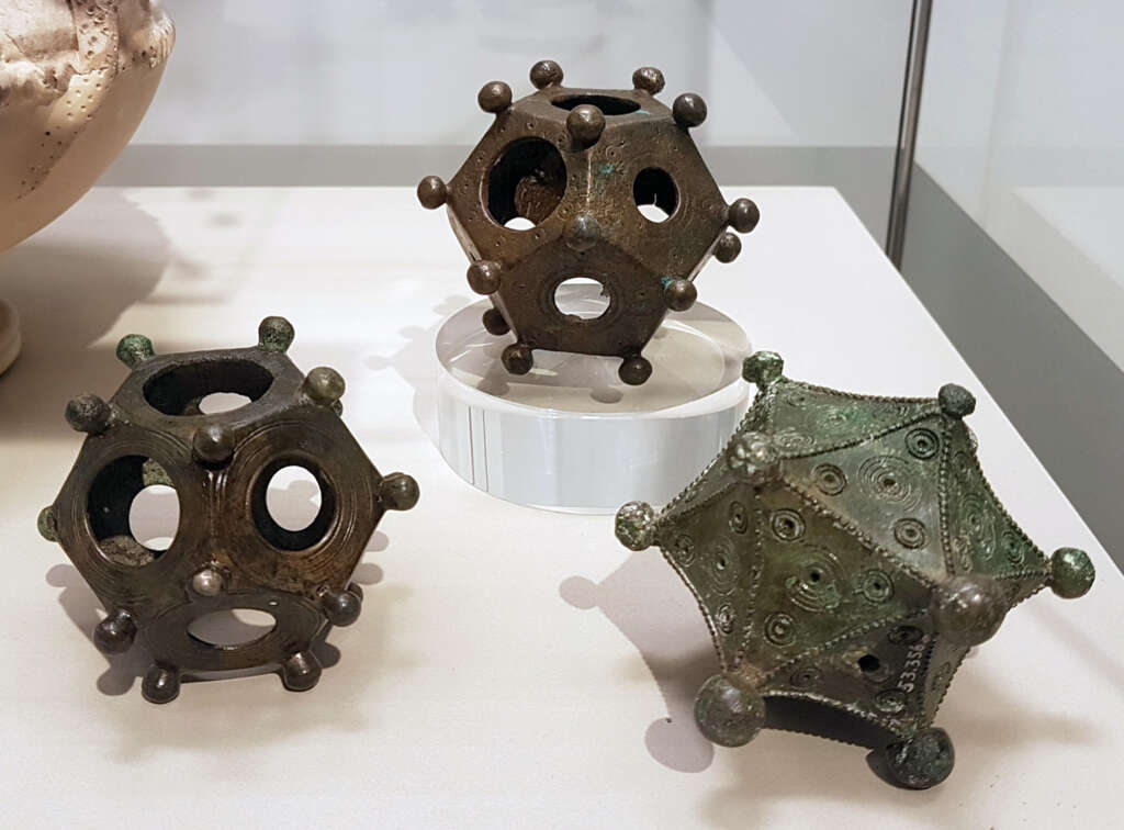 Three ancient Roman dodecahedrons made of metal are displayed on a surface. They have circular holes on each face and protruding knobs at each vertex. One is elevated on a white stand.