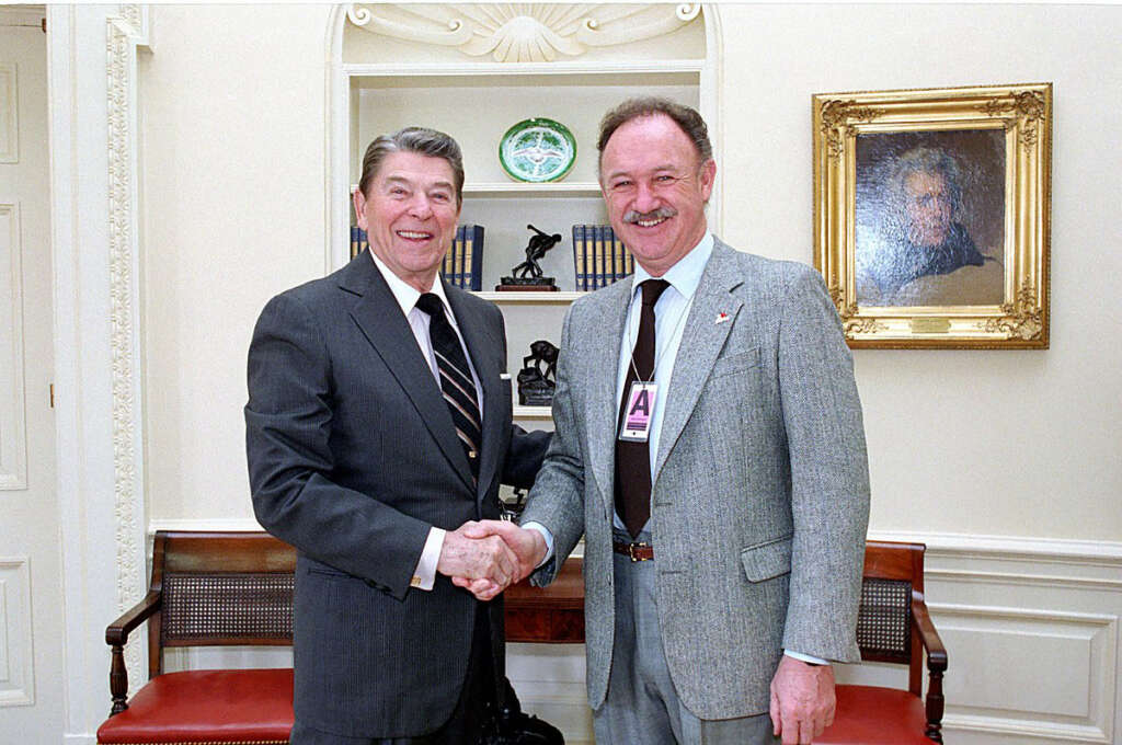 Two men in suits shake hands in a room with a portrait on the wall and a decorative shelf. One man has gray hair, and the other has a mustache. The setting appears formal, with elegant wood furniture and decor.