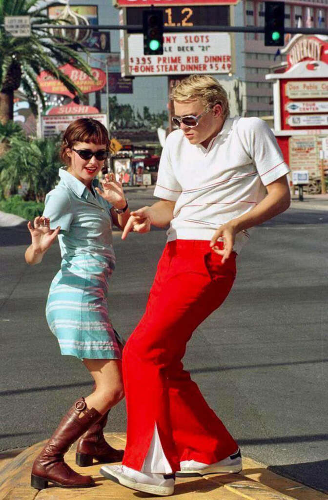 Two people in retro outfits dance joyfully on a street. The person on the left wears a blue dress and brown boots, while the person on the right sports red bell-bottom pants and a white shirt. A vintage diner sign and palm trees are visible in the background.