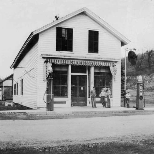 A vintage black-and-white photo of a small gas station with a wooden facade and large windows. Two men and a child stand outside. A gas pump is visible in the foreground. The area is slightly hilly with trees in the background.