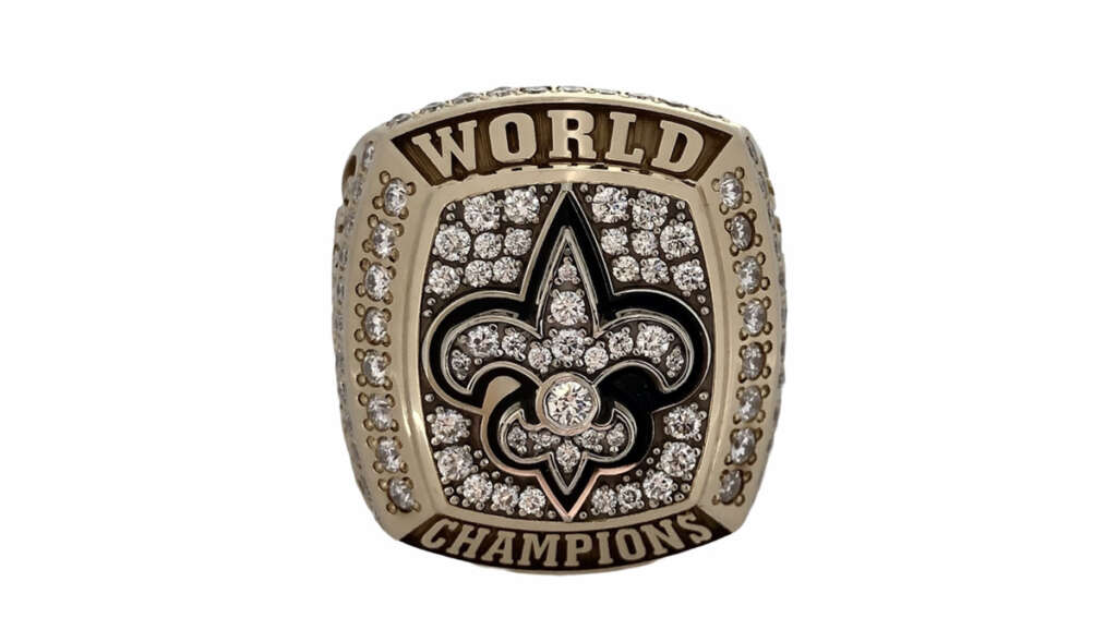 A gold and diamond ring with "World Champions" engraved on it. The center features a large fleur-de-lis symbol encrusted with diamonds, surrounded by additional small diamonds.