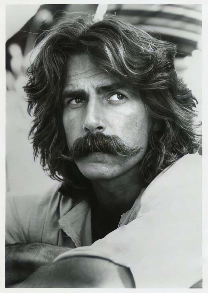 A person with long, wavy hair and a thick mustache gazes thoughtfully to the side. The black and white photo highlights their serious expression and detailed features. They rest their arm casually, adding to the contemplative mood.